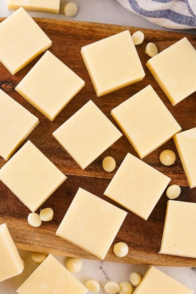 Neatly cut white chocolate squares on a wooden board, surrounded by scattered chips on a marble surface with striped cloth.