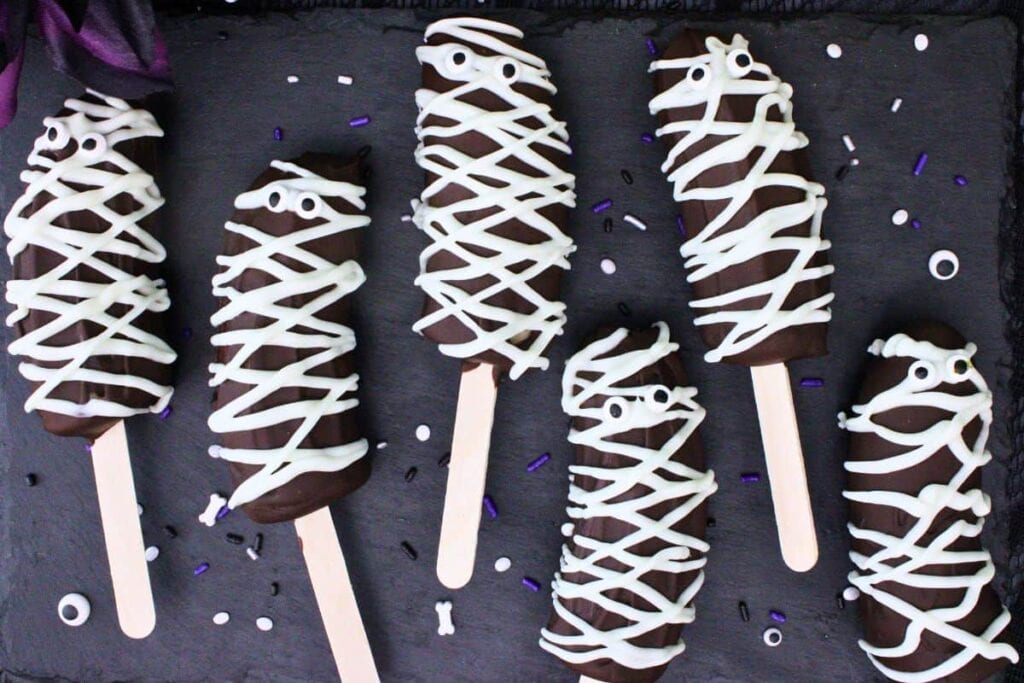 Six chocolate-covered frozen banana pops are decorated as Halloween mummies with white icing wrapped across each banana and candy eyes peeking through. The mummy banana pops are arranged on a black serving board sprinkled with Halloween-themed sprinkles, creating a fun and spooky dessert.