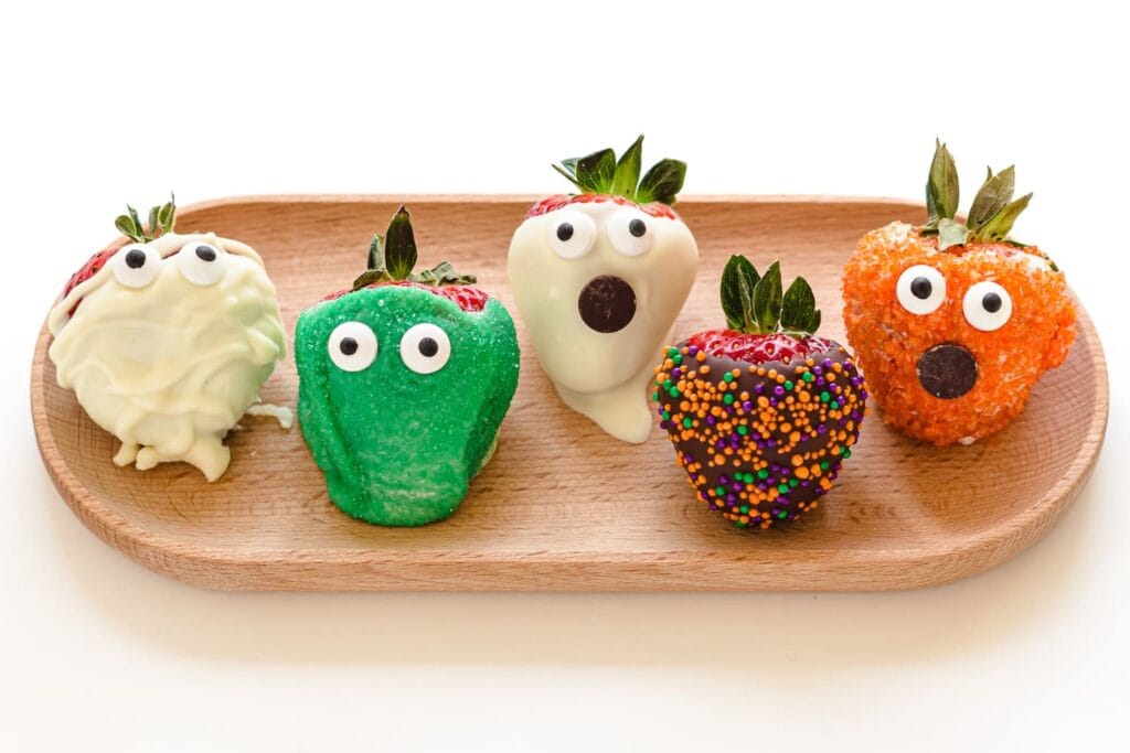 A wooden serving tray holds five Halloween-themed chocolate-covered strawberries decorated as spooky characters. The assortment includes white chocolate mummy and ghost strawberries, a green monster strawberry, a chocolate-covered strawberry with orange and purple sprinkles, and an orange-coated monster strawberry. Candy eyes and chocolate mouths give each strawberry a playful Halloween expression.