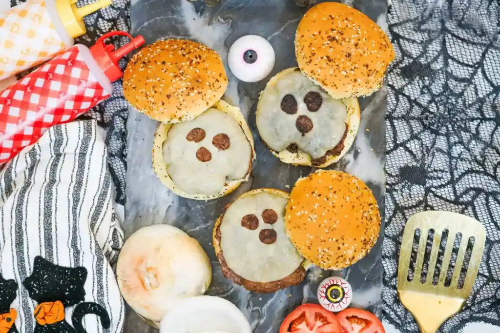 Three Halloween-themed cheeseburgers are arranged on a dark serving board, each topped with melted white cheese cut into ghost faces and decorated with olive slices for the eyes and mouth. The burgers are surrounded by burger buns, sliced onions and tomatoes, condiment bottles, candy eyeballs, and spooky black lace decorations.