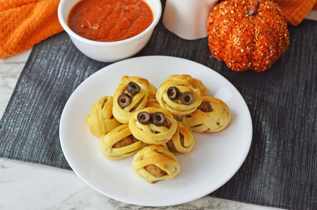 Halloween mummy sausage bites wrapped in golden pastry with olive eyes, served on a white plate with marinara dipping sauce.