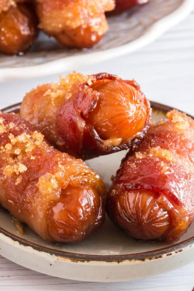 Ceramic plate of bacon-wrapped sausages glazed with caramelized brown sugar, crispy and golden, with more pieces visible in background.