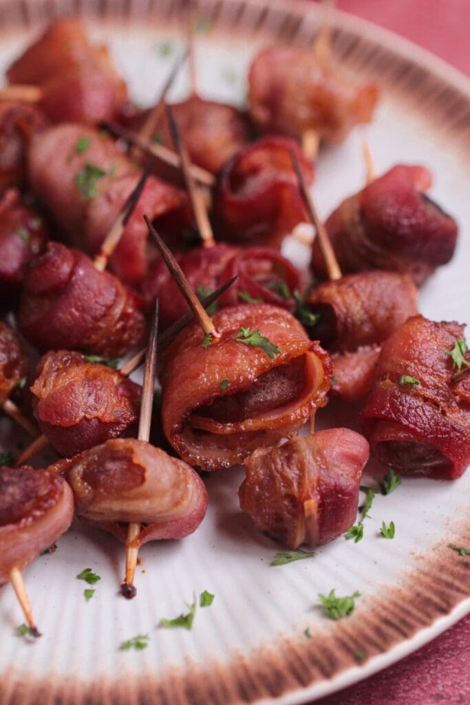 Plate of crispy bacon-wrapped bites secured with toothpicks, garnished with parsley, on a decorative rimmed plate over a reddish surface.