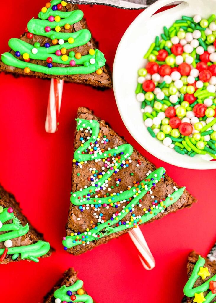 Triangular brownies decorated as Christmas trees with green frosting zigzags, colorful sprinkles, and candy cane trunks, set on bright red background with bowl of sprinkles nearby.