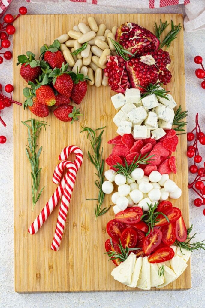 Wooden board with strawberries, pomegranate, cheeses, mozzarella balls, cherry tomatoes, and candy canes decorated with rosemary sprigs.