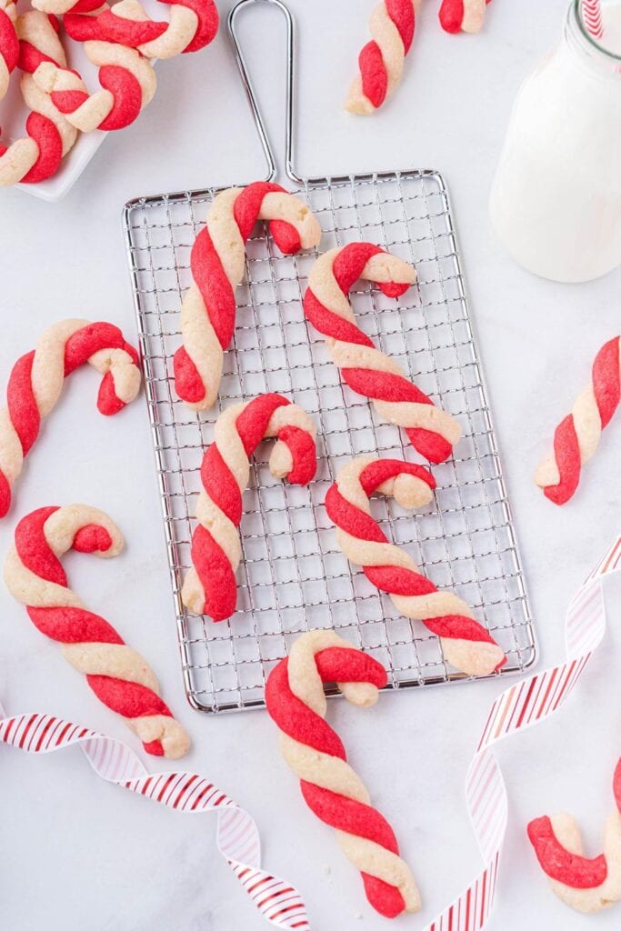 Twisted red-and-white candy cane cookies on cooling rack with milk bottle and striped straw, accented by festive ribbon.