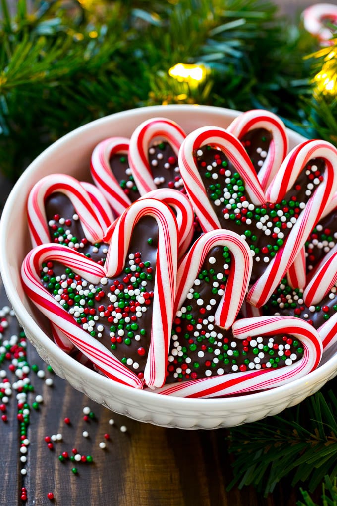 Candy cane hearts filled with chocolate and topped with festive sprinkles, arranged in white bowl with evergreen branches and lights.