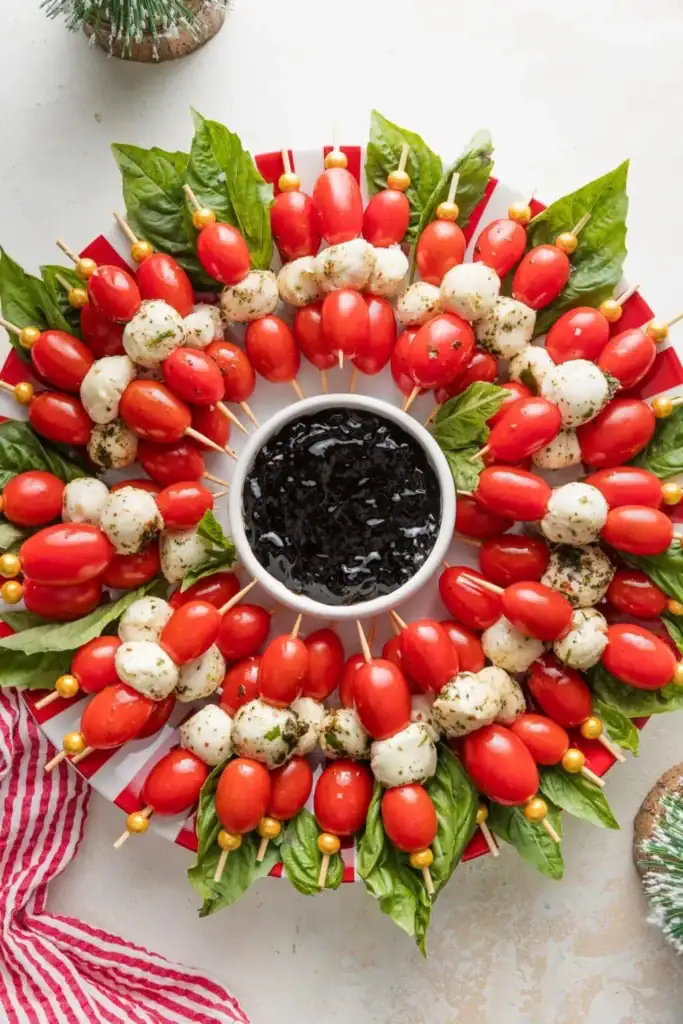 Circular wreath of skewered grape tomatoes, mozzarella, and basil around a bowl of balsamic glaze, decorated with gold beads on a festive red-and-white plate.