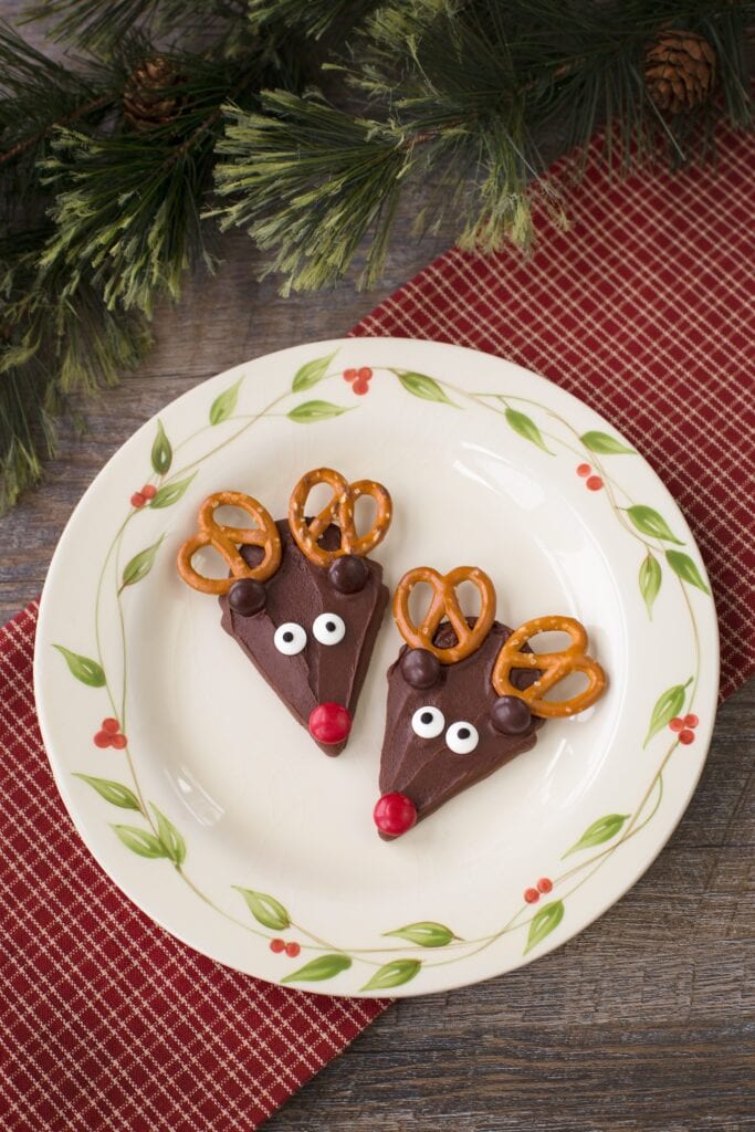 Triangular chocolate treats decorated as reindeer with pretzel antlers, candy eyes, ears, and red noses on festive plate with pine branches and checkered cloth.