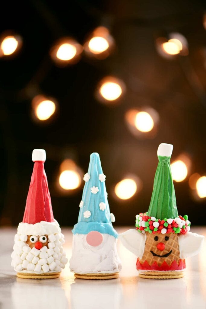 Festive cone desserts decorated as Santa, gnome, and elf with frosting hats, candy faces, and marshmallow beards, set against warm holiday lights.