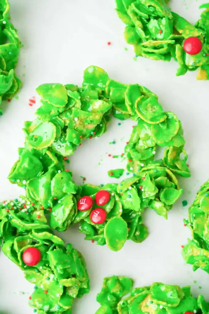 Green cornflake wreath cookies decorated with sprinkles and red candy berries, arranged on white surface.