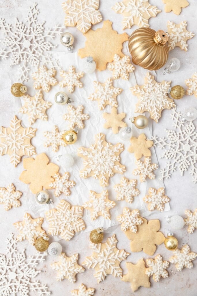 Snowflake-shaped cookies decorated with white icing and powdered sugar, arranged on light surface with gold, silver, and white ornaments.