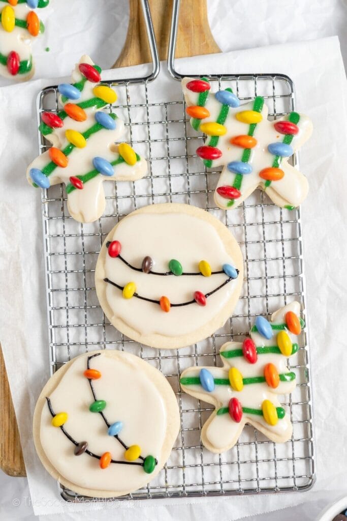Sugar cookies shaped as trees and circles decorated with icing and colorful candy lights on cooling rack with parchment paper.