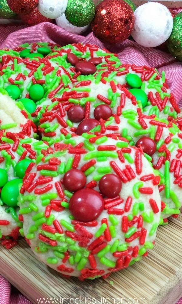 Cookies with red and green sprinkles and candy pieces on wooden surface with pink cloth and glittery ornaments.