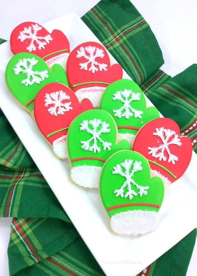 Mitten-shaped cookies decorated with red and green icing, white snowflakes, and sugar-coated cuffs, arranged on white plate over green plaid cloth.