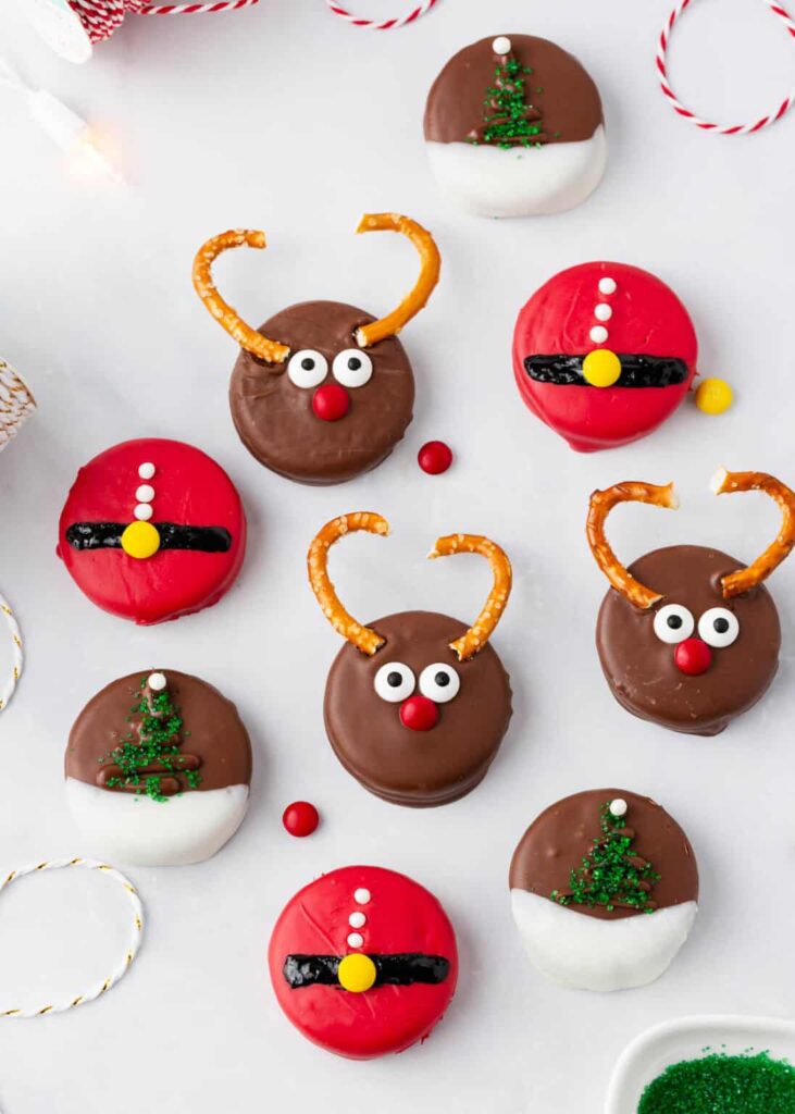 Assorted chocolate-covered cookies decorated as reindeer, Santa belts, and Christmas trees, arranged on white surface with red candies, pretzels, and holiday string.