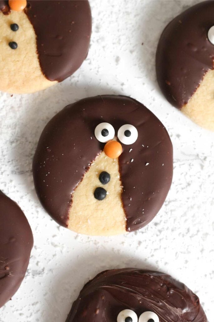 Round cookies dipped in chocolate, decorated as penguins with candy eyes and orange noses, arranged on white textured surface.