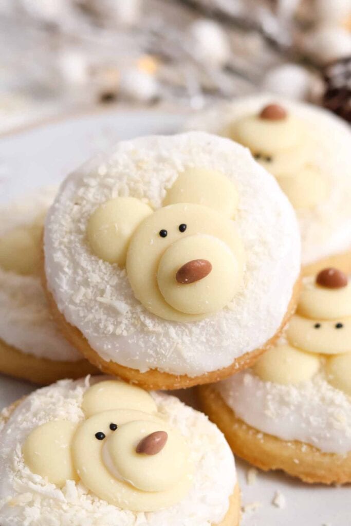 White-iced cookies decorated as polar bear faces with chocolate ears, candy eyes, and brown noses, arranged on plate with snowy flakes.