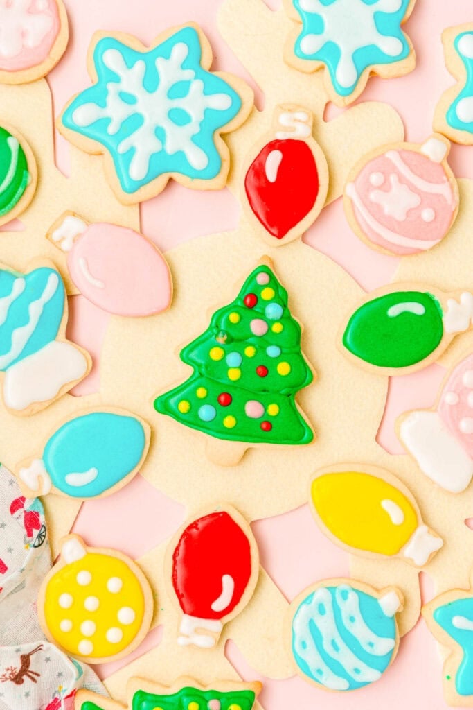 Assorted Christmas sugar cookies shaped like trees, ornaments, and snowflakes, decorated with colorful icing and sprinkles, arranged on pink background.