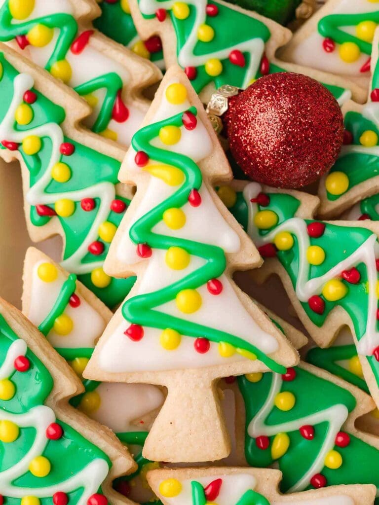 Christmas tree-shaped sugar cookies decorated with green, white, red, and yellow icing, arranged with a red glitter ornament on festive background.