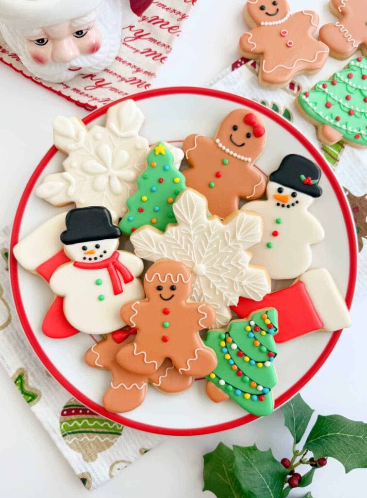 Plate of holiday cookies shaped as gingerbread people, snowflakes, trees, snowmen, and stockings, decorated with colorful icing and candy details, set on festive cloth with ceramic Santa in background.