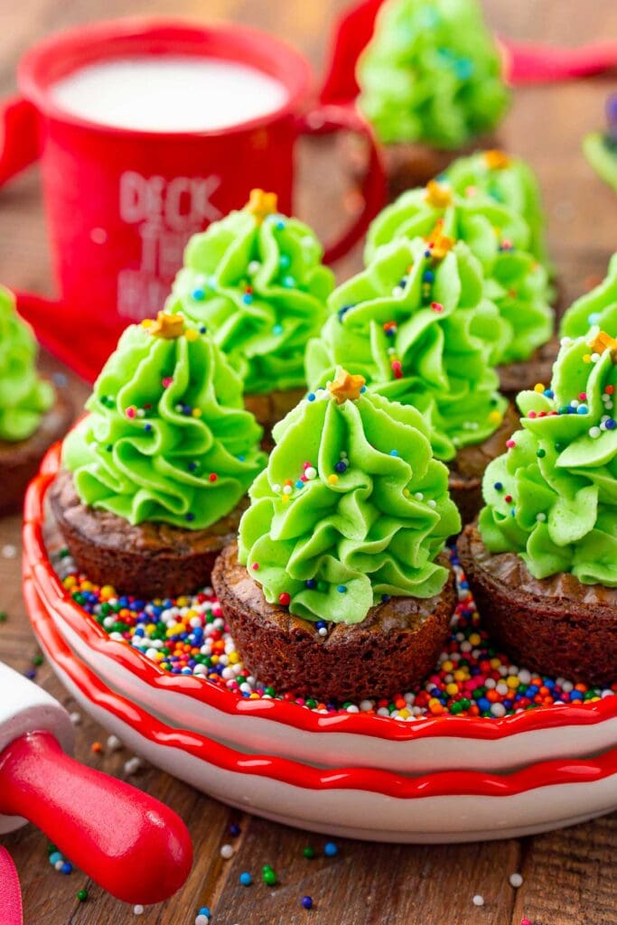 Brownie cups topped with green frosting Christmas trees decorated with sprinkles and star candies, arranged on red dish of sprinkles beside festive mug of milk.