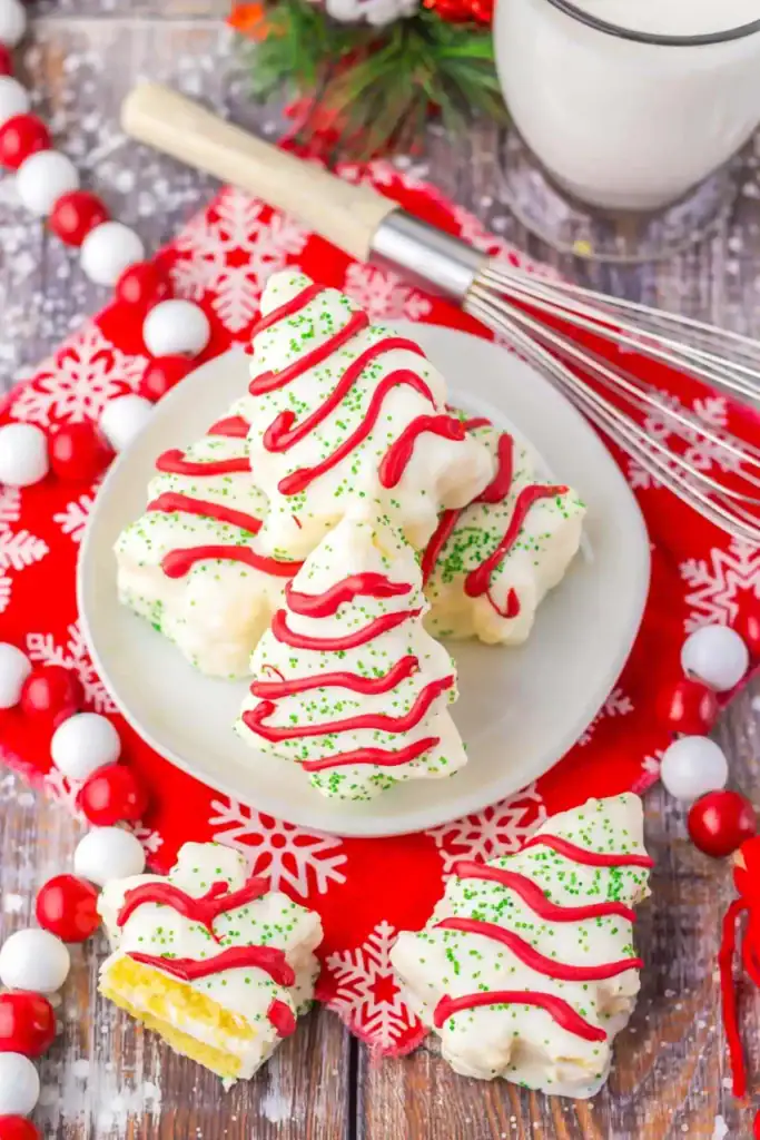 White plate of Christmas tree-shaped iced cakes with red drizzle and green sprinkles, one cut open showing yellow cake and white filling, set on snowflake napkin with festive decorations and milk nearby.