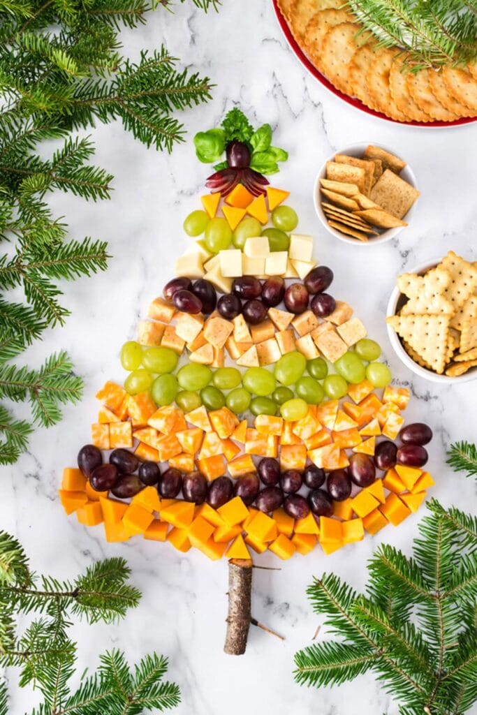 Festive Christmas tree made of cheese cubes, grapes, and crackers, topped with leafy greens, set on wood with evergreen branches and bowls of crackers nearby.