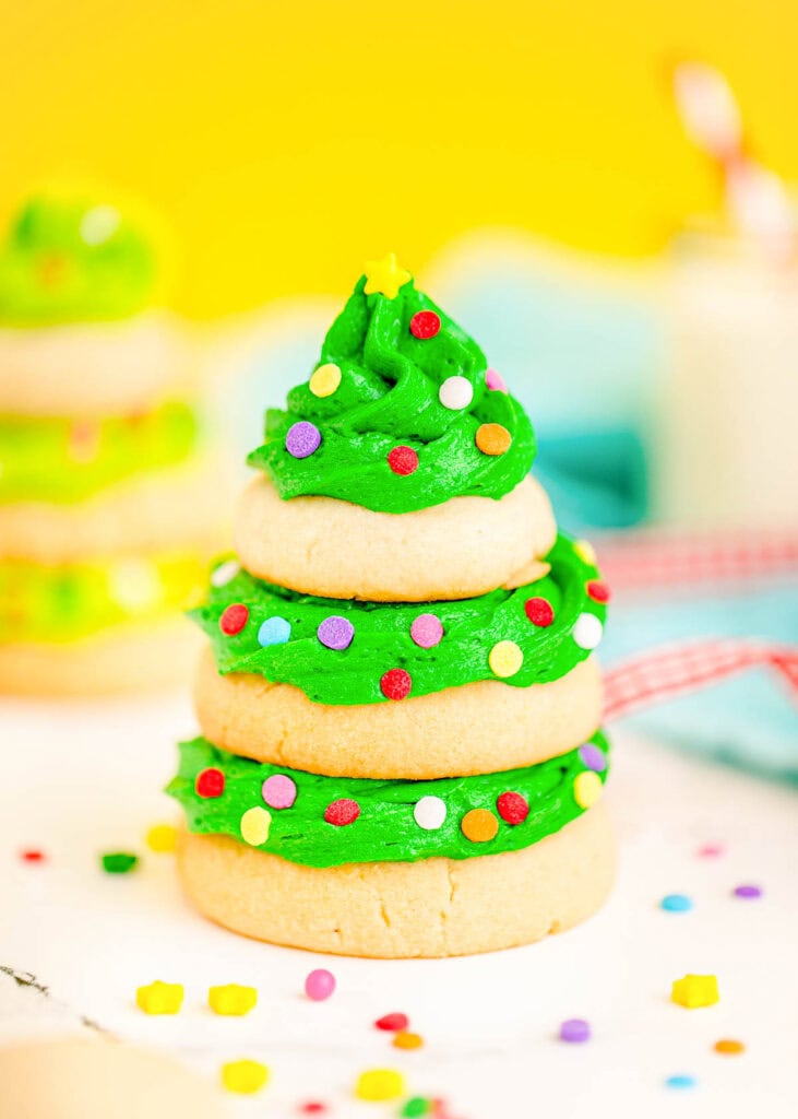 Stacked sugar cookies with green frosting layers shaped like Christmas tree, decorated with sprinkles and star topper, set against bright yellow background with milk and festive accents.