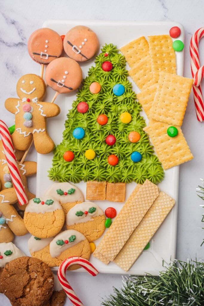 Festive platter with frosting Christmas tree centerpiece, surrounded by gingerbread cookies, dipped round cookies, wafers, crackers, caramel squares, candy canes, and Santa-themed lollipops.