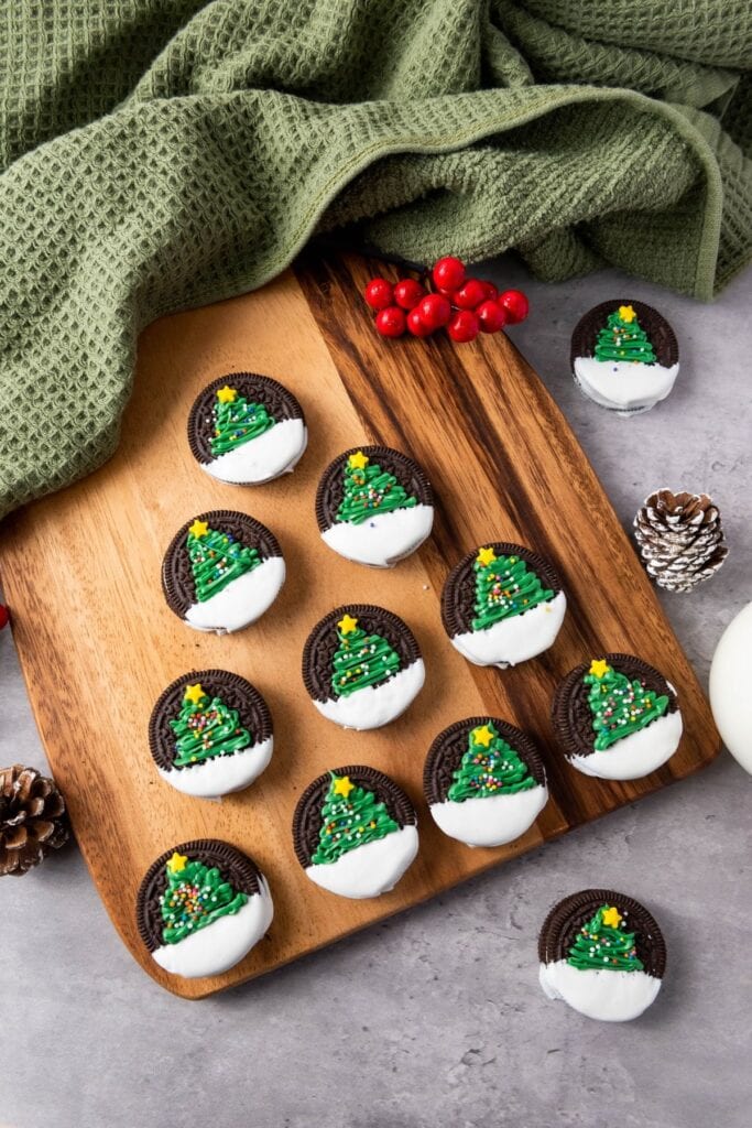 Chocolate sandwich cookies half-dipped in icing, topped with green frosting Christmas trees decorated with sprinkles and yellow stars, arranged on wooden board with festive berries and pinecones.