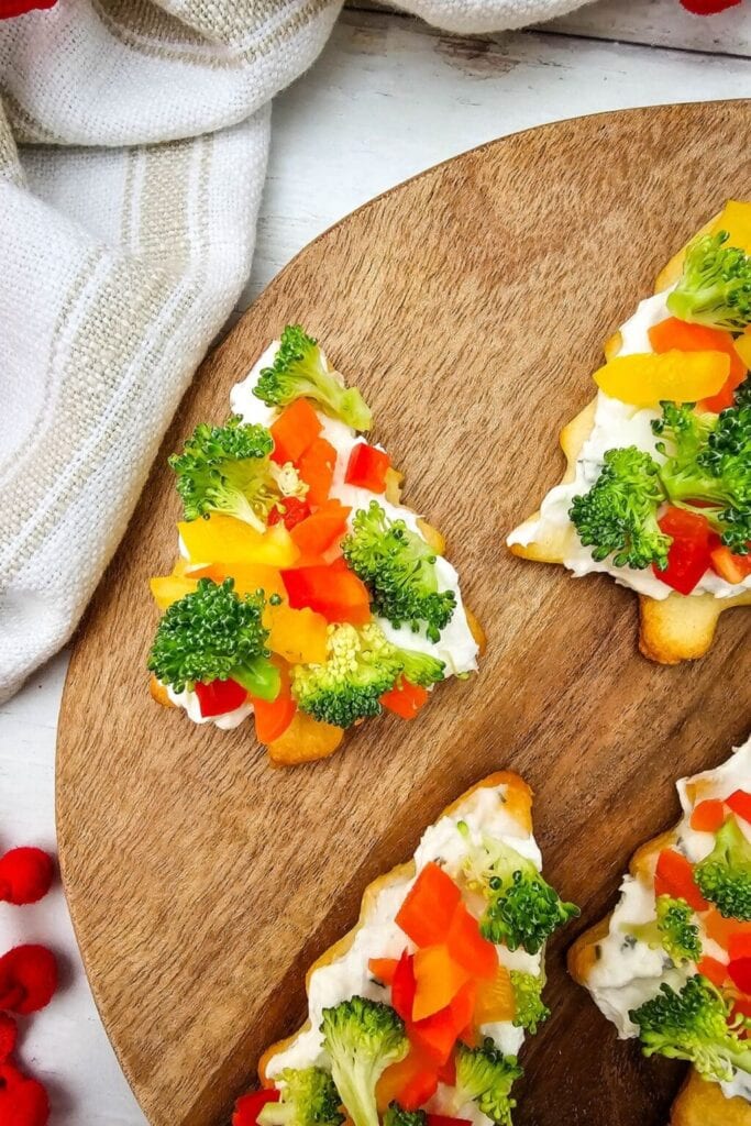 Wooden board with Christmas tree-shaped pastry bases topped with creamy spread, decorated with broccoli florets and colorful bell pepper pieces like ornaments.