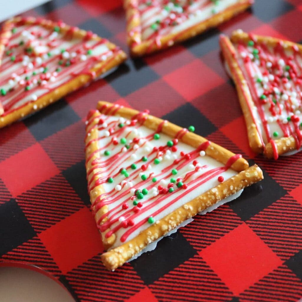 Triangular pretzel treats filled with white icing, drizzled with red, and topped with festive sprinkles, arranged on red plaid surface.