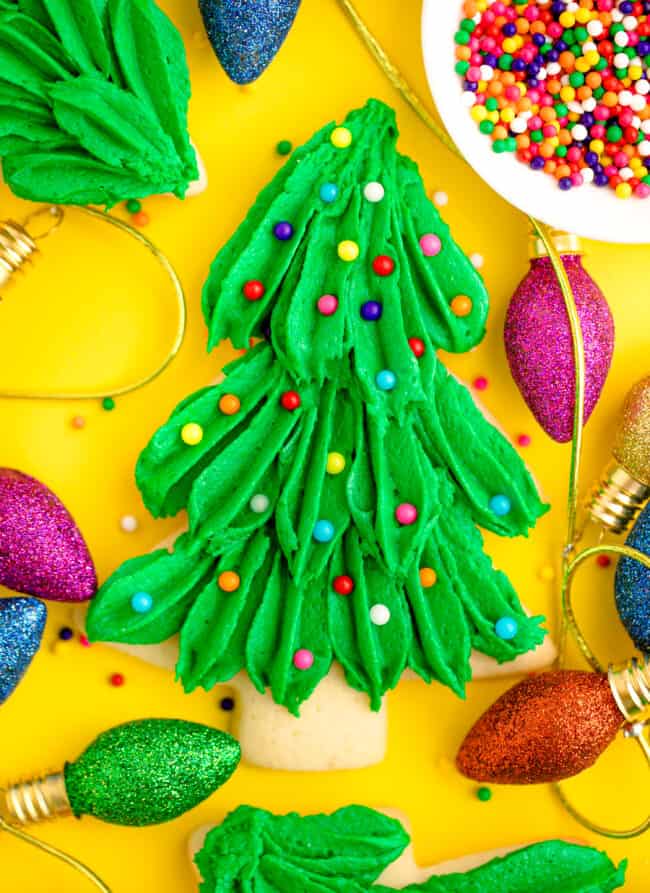 Christmas tree-shaped cookie with green frosting and colorful sprinkles, placed on yellow background with glittery light decorations and dish of sprinkles.