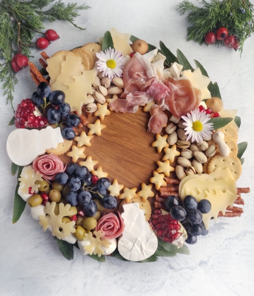 Circular charcuterie wreath of cheese cutouts, cured meats, olives, blueberries, pomegranate, pistachios, crackers, and pretzels with decorative flowers and leaves on wood.
