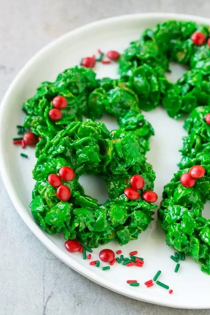 Green cornflake wreath cookies decorated with red candy berries, arranged on white plate with festive sprinkles.