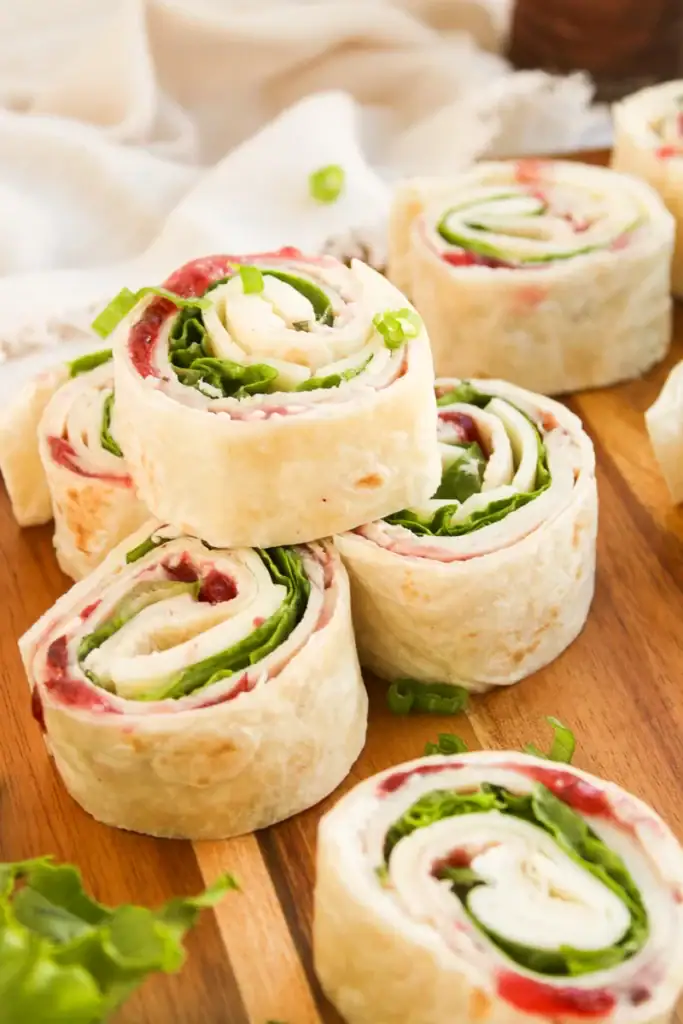 Wooden surface with sliced tortilla pinwheels filled with turkey, lettuce, cheese, and cranberry spread, showing colorful spiral layers.