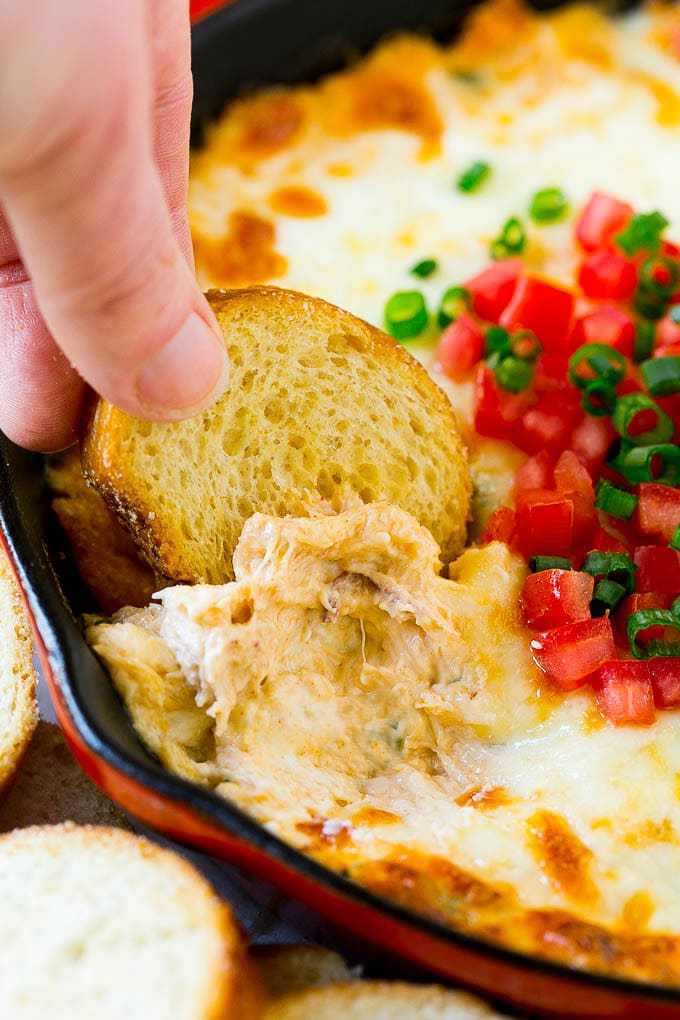 Red baking dish of cheesy dip topped with tomatoes and green onions, with toasted bread slices and a hand dipping crostini into the melted mixture.