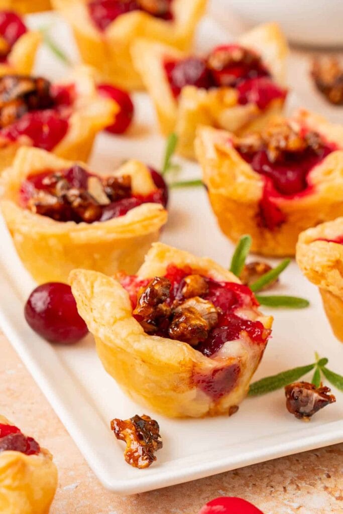 Golden pastry cups filled with cranberry sauce and candied nuts, arranged on a white plate with fresh cranberries and rosemary sprigs.