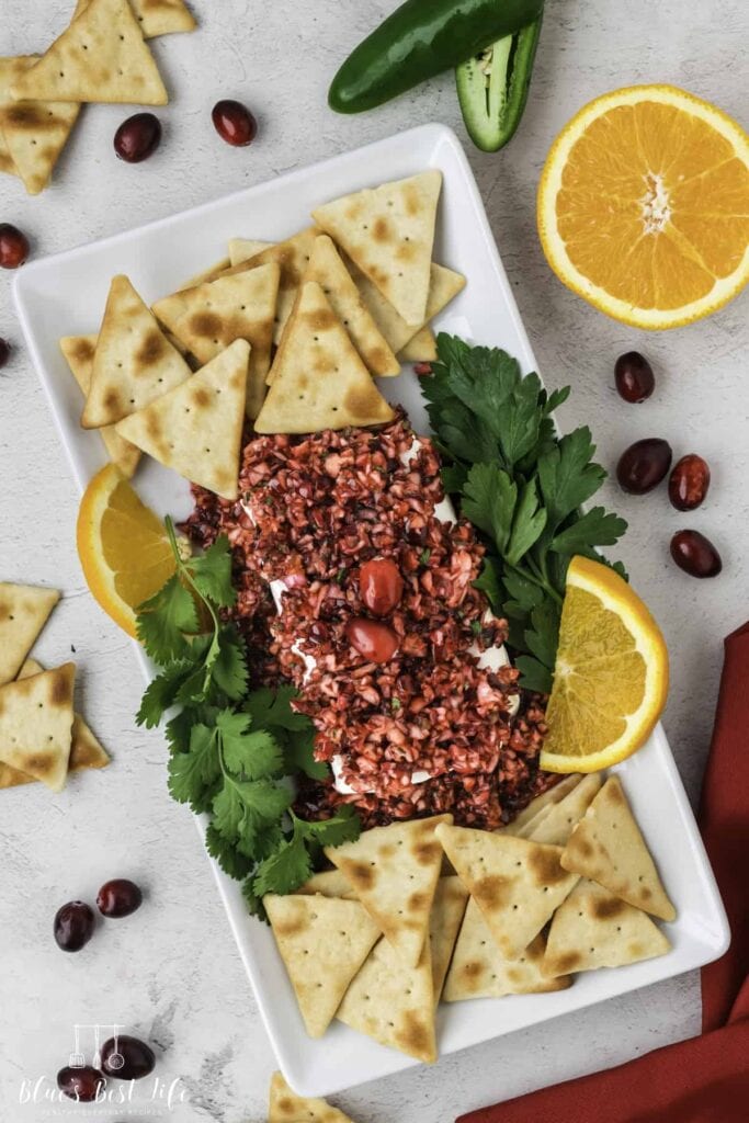 White dish with cream cheese topped by cranberry relish, surrounded by crackers, orange slices, parsley, cranberries, and jalapeño on a light surface.