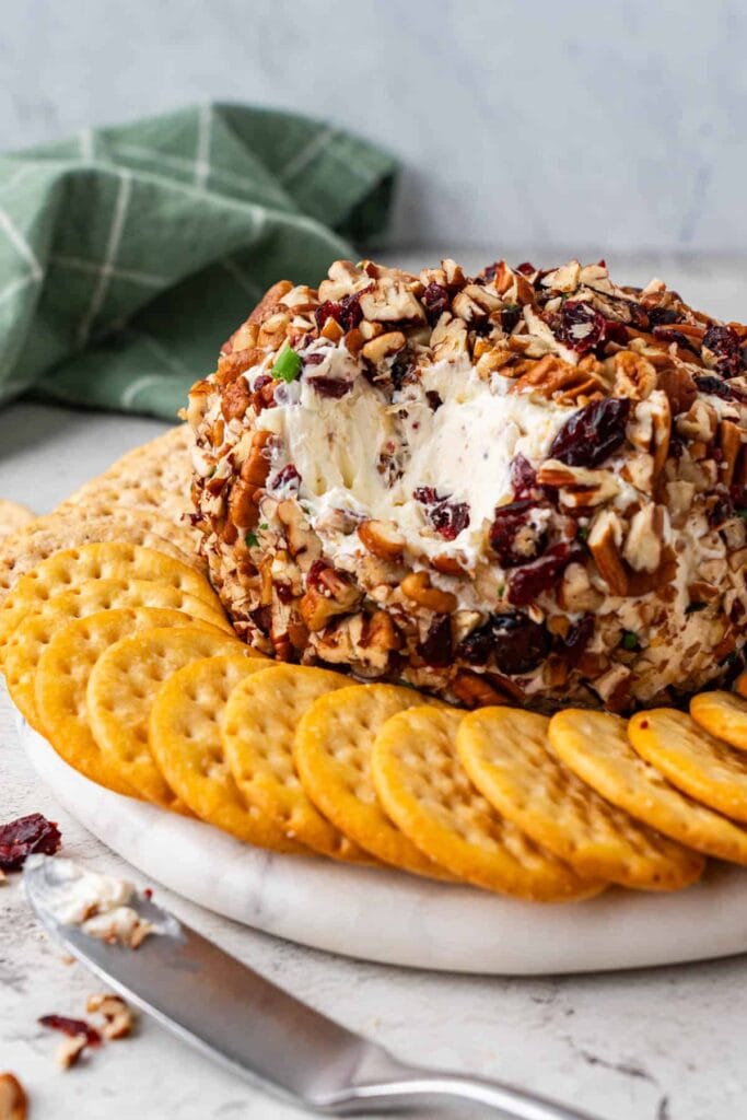 Nut- and cranberry-coated cheese ball with creamy interior, served on a white platter surrounded by crackers, with spreading knife and green checkered cloth.