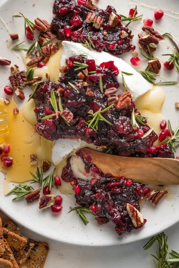 Baked brie topped with cranberry-pomegranate sauce, garnished with rosemary, pecans, and honey drizzle, served with crackers on a white plate.
