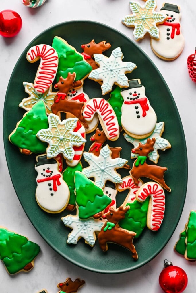 Festive sugar cookies shaped as snowmen, reindeer, trees, candy canes, and snowflakes, decorated with colorful icing and arranged on dark green oval plate with ornaments.