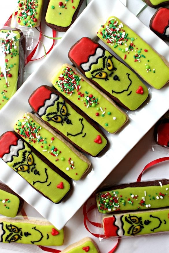 Rectangular cookies decorated as the Grinch with green icing, Santa hat detail, and sprinkles, arranged on white plate with some wrapped in clear plastic tied with red ribbon.