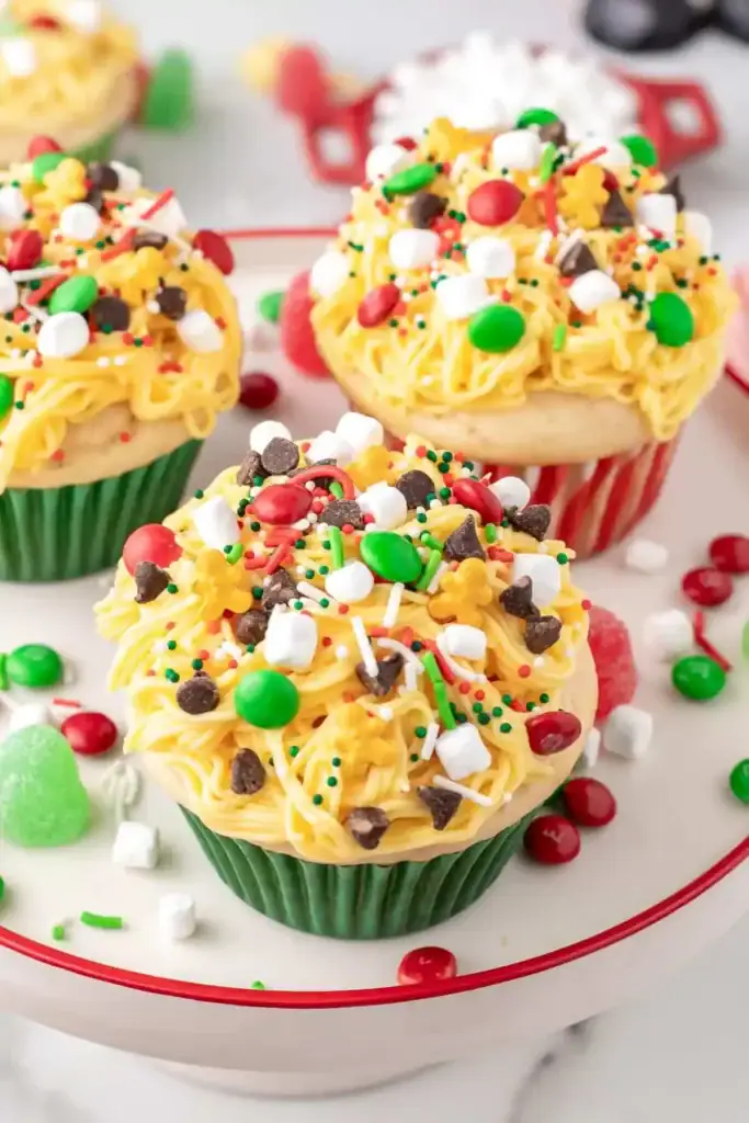 Cupcakes with yellow frosting swirls topped with sprinkles, marshmallows, chocolate chips, and red-green candies in holiday liners, arranged on white plate with red rim.