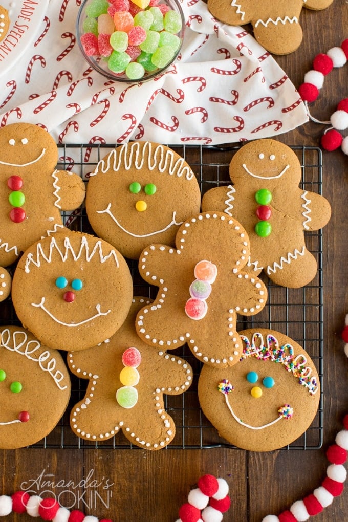 Gingerbread cookies decorated with icing and colorful candies, shaped as people and round faces, arranged on cooling rack with pom-pom garland and gumdrop bowl.