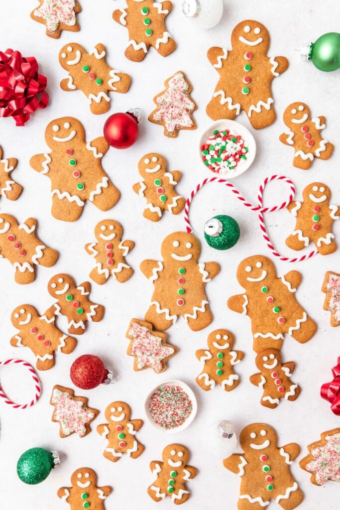 Gingerbread cookies shaped as people and trees, decorated with icing and candy buttons, arranged with ornaments, sprinkles, ribbons, and striped string for festive Christmas atmosphere.