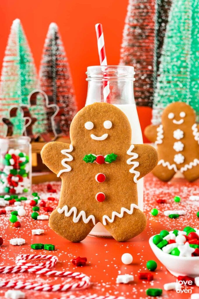 Gingerbread cookie with icing and holly decoration leaning against milk bottle with striped straw, surrounded by festive trees, candies, and powdered sugar sprinkles.