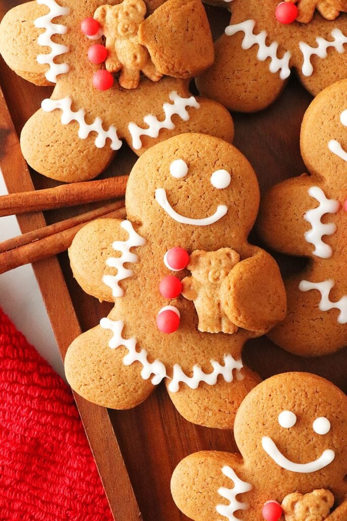 Gingerbread people decorated with icing smiles and red candy buttons, each holding a small bear-shaped cookie, arranged on wooden tray with cinnamon sticks.