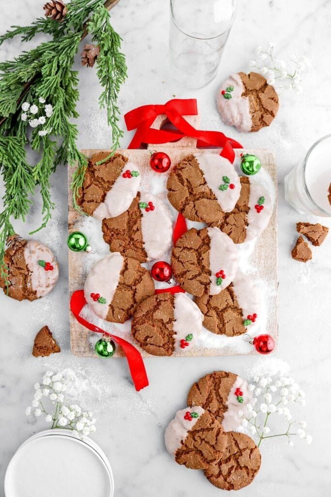 Brown cracked cookies half-dipped in white icing, decorated with holly sprinkles, arranged on wooden board with powdered sugar, ornaments, greenery, and milk glasses.